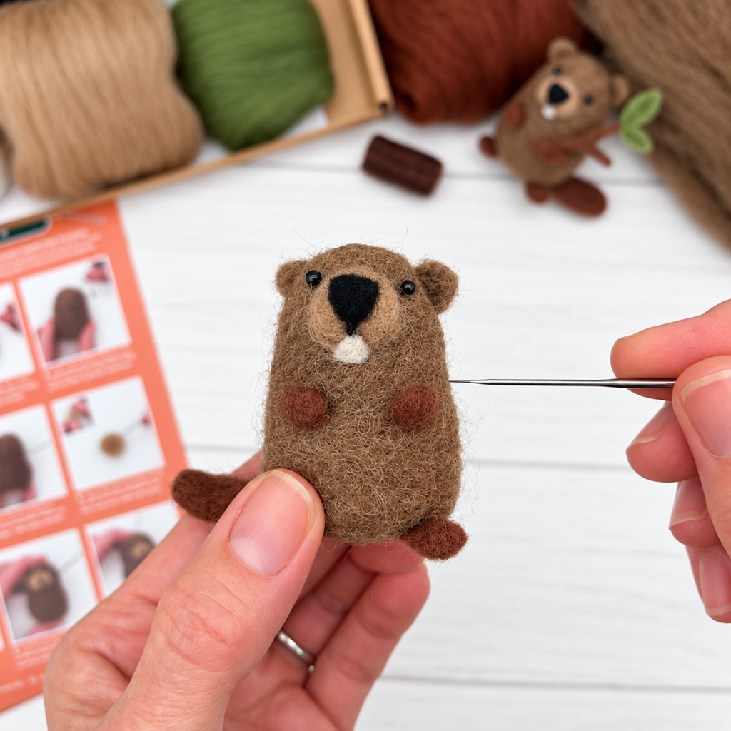 Close-up of hands holding a partially completed brown wool beaver while adding details with a felting needle, with illustrated instructions and wool fibres in the background.