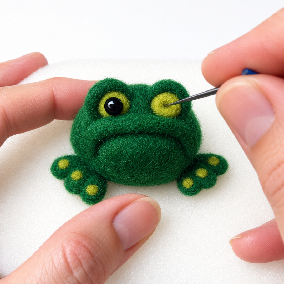 Close-up of hands holding a dark green needle felted frog with raised yellow eyes, a smiling mouth and little green feet.