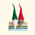 Two handmade Scandinavian wool gnomes with tall red and green pointed hats, long white beards and round noses, displayed on a stack of colourful books.