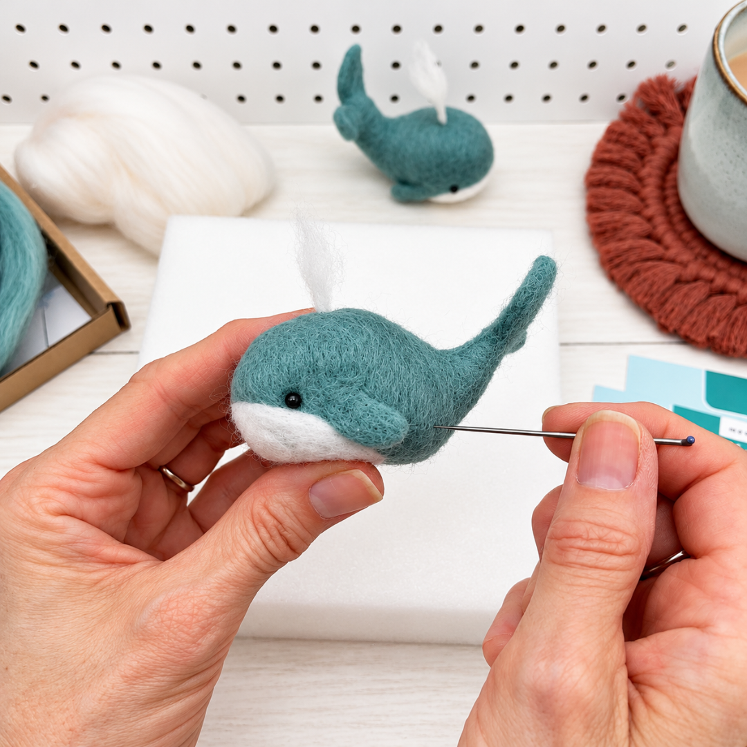 Close-up of a handmade blue wool whale held between fingers, showing the finished design from the Bergin And Bath needle felting kit.