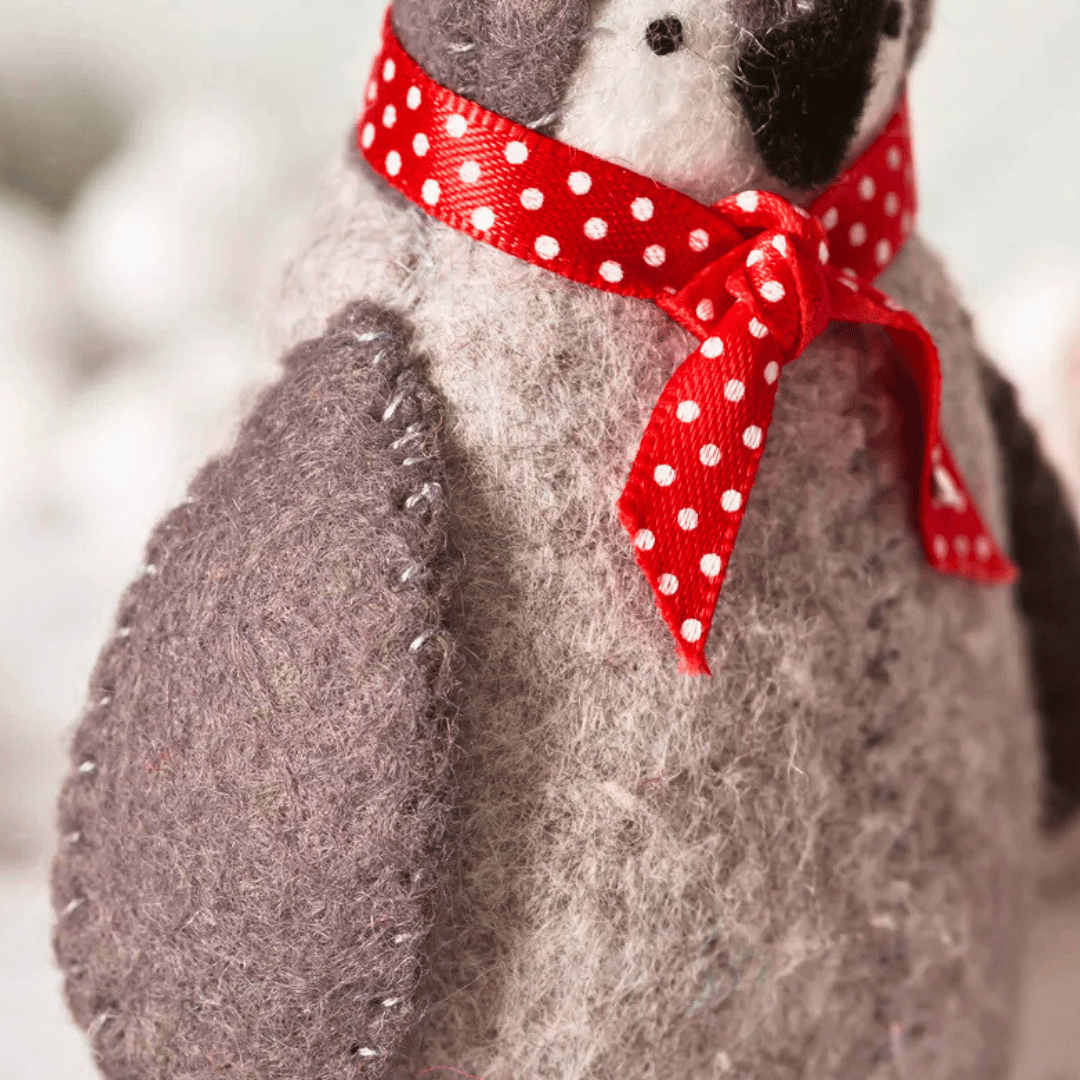 A close-up of a felt penguin toy wearing a red ribbon bow with white polka dots, set against a soft, blurred white background.