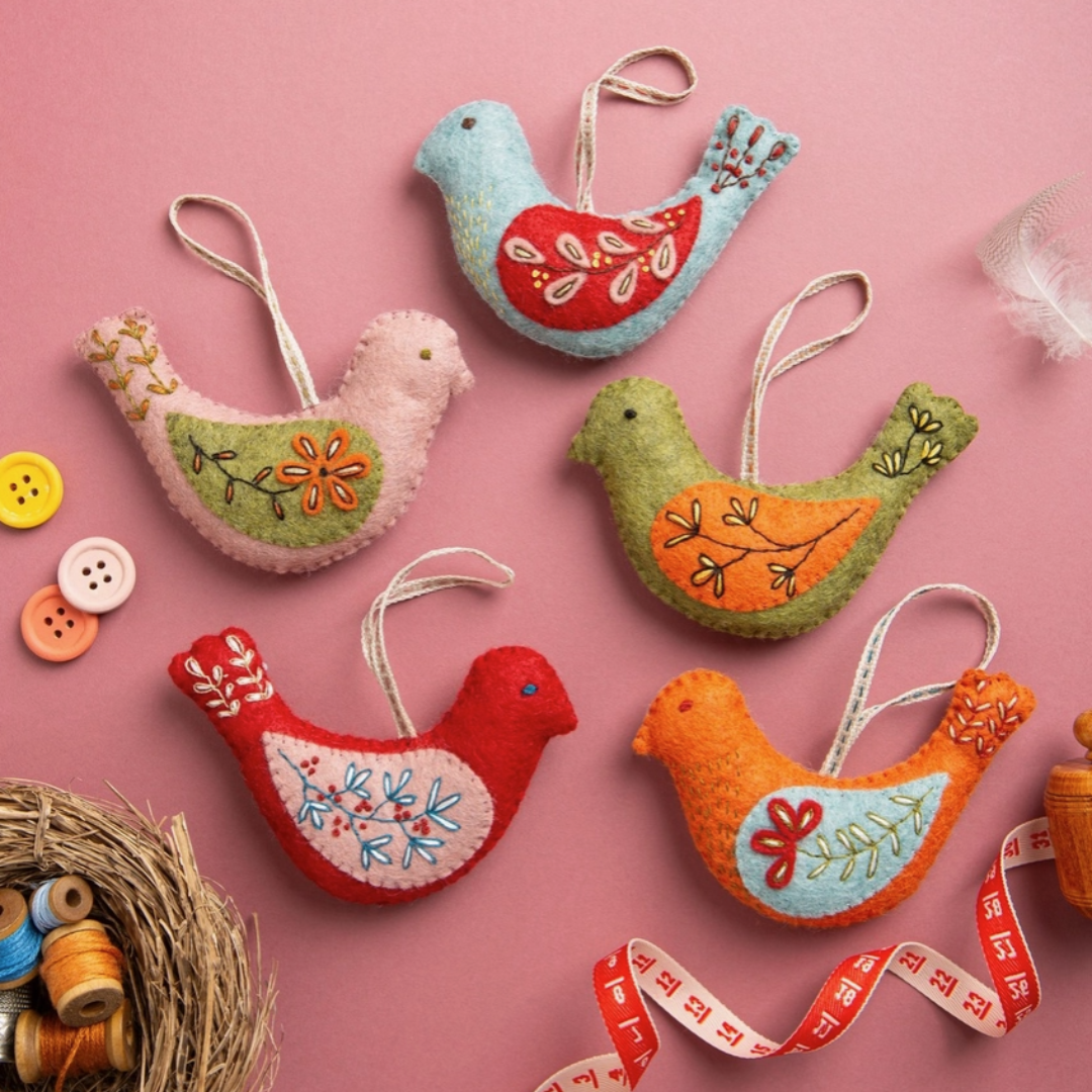Set Of 5 Folk Birds Felt Craft Kit by Corinne Lapierre with completed bird ornaments, buttons, ribbon and crafting accessories displayed on a pink background.