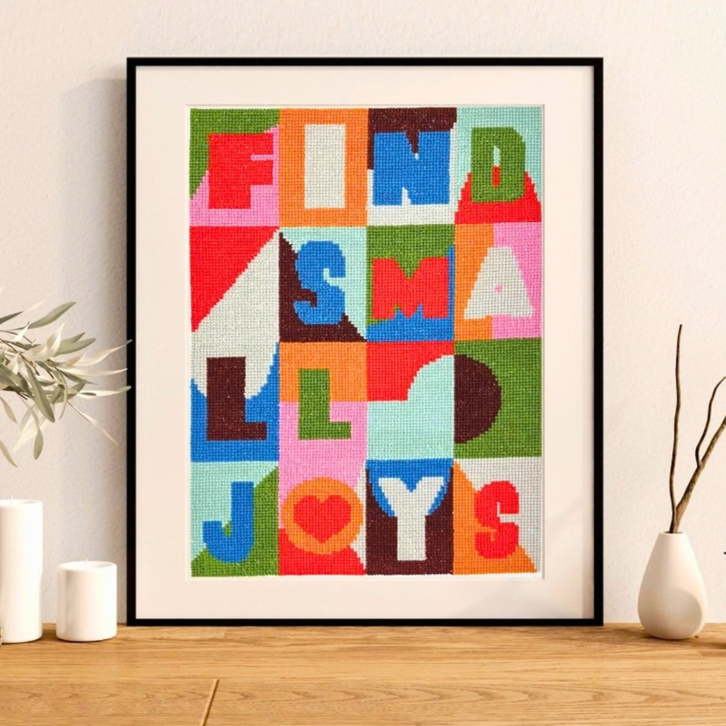 Small Joys cross stitch by Budgiegoods displayed in a black frame on a styled timber shelf.