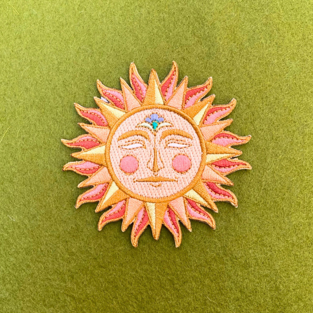Close-up of Ashlea Bechaz Illustration sun embroidered iron on patch featuring a smiling celestial face and vibrant pink, orange and yellow stitching against a green background.