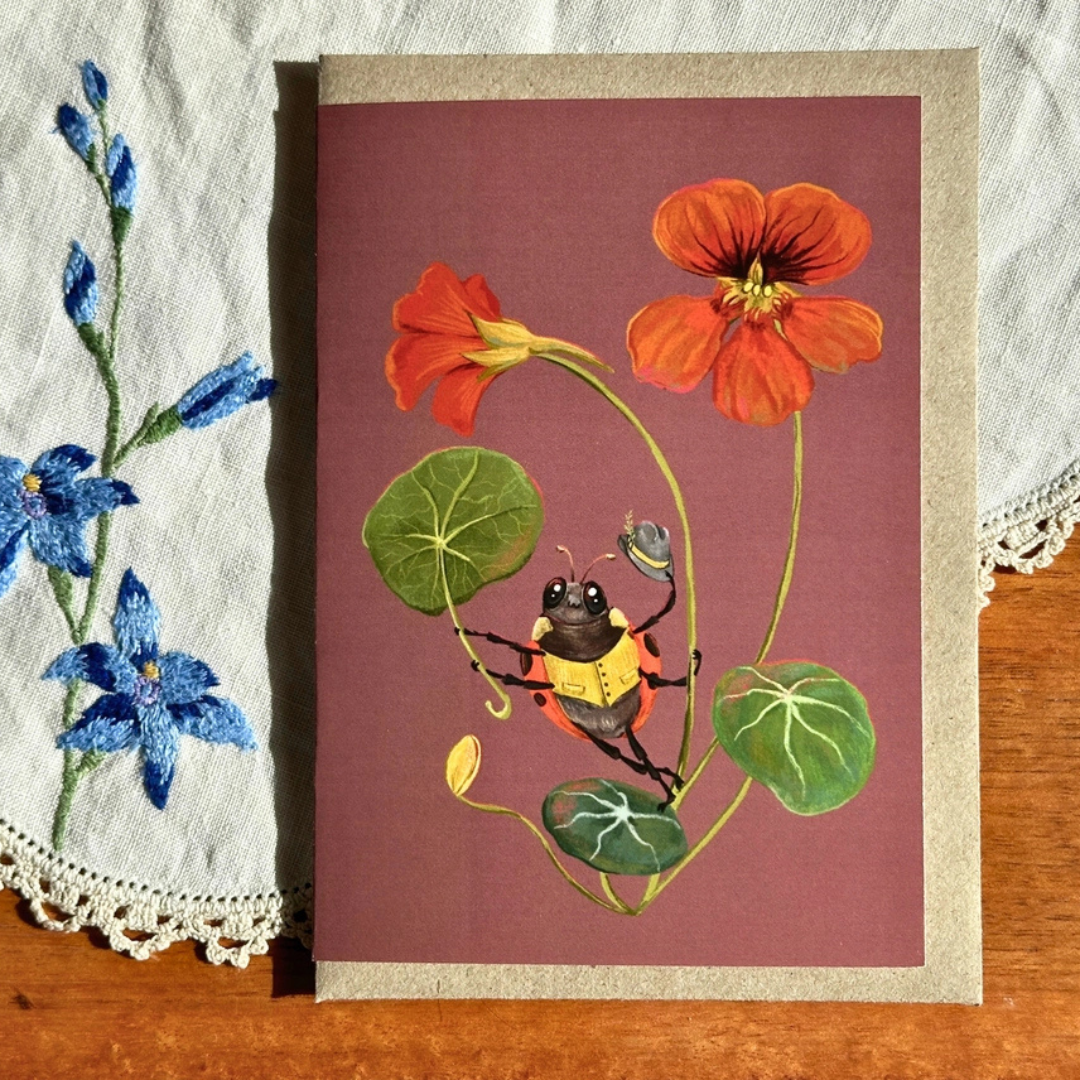 Sun Shower greeting card styled on a wooden surface with lace and blue flowers, featuring a colourful bee and nasturtium illustration.