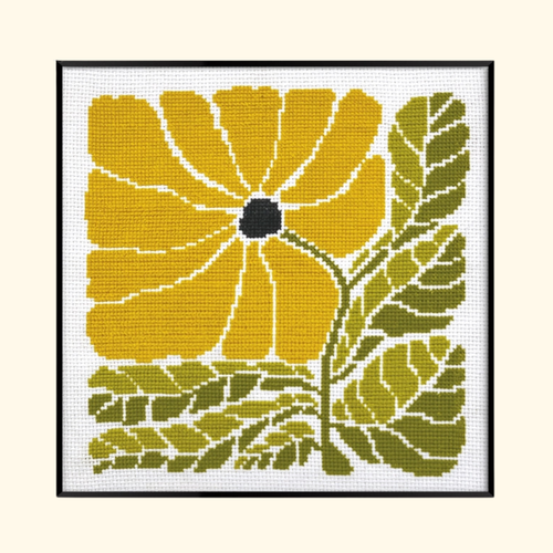 A framed cross-stitch or needlepoint floral artwork featuring a large yellow flower with a dark center and olive green leaves on a white stitched background, shown against a light beige backdrop.
