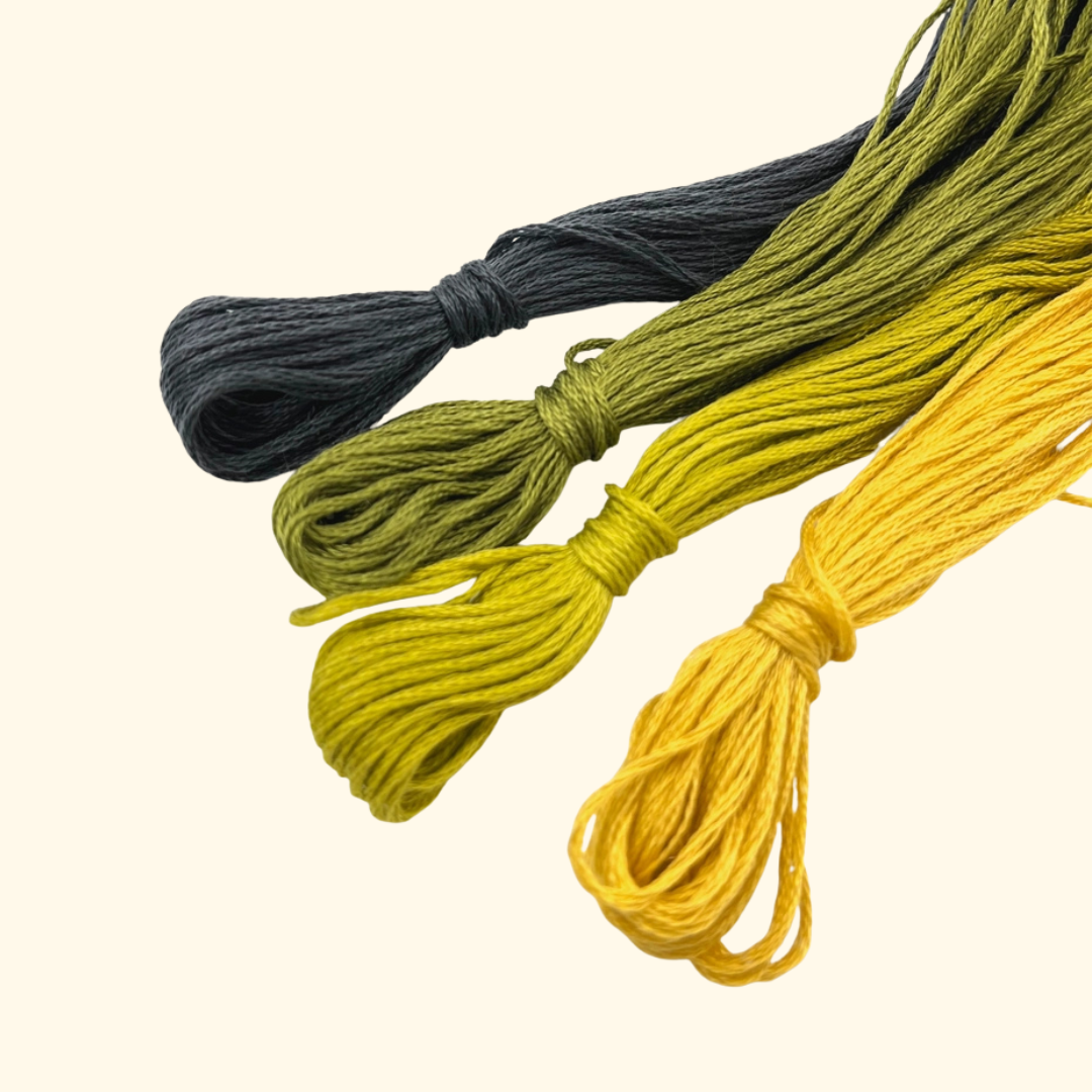 Three coiled bundles of embroidery floss in dark charcoal gray, olive green, and golden yellow, arranged diagonally on a light cream background.