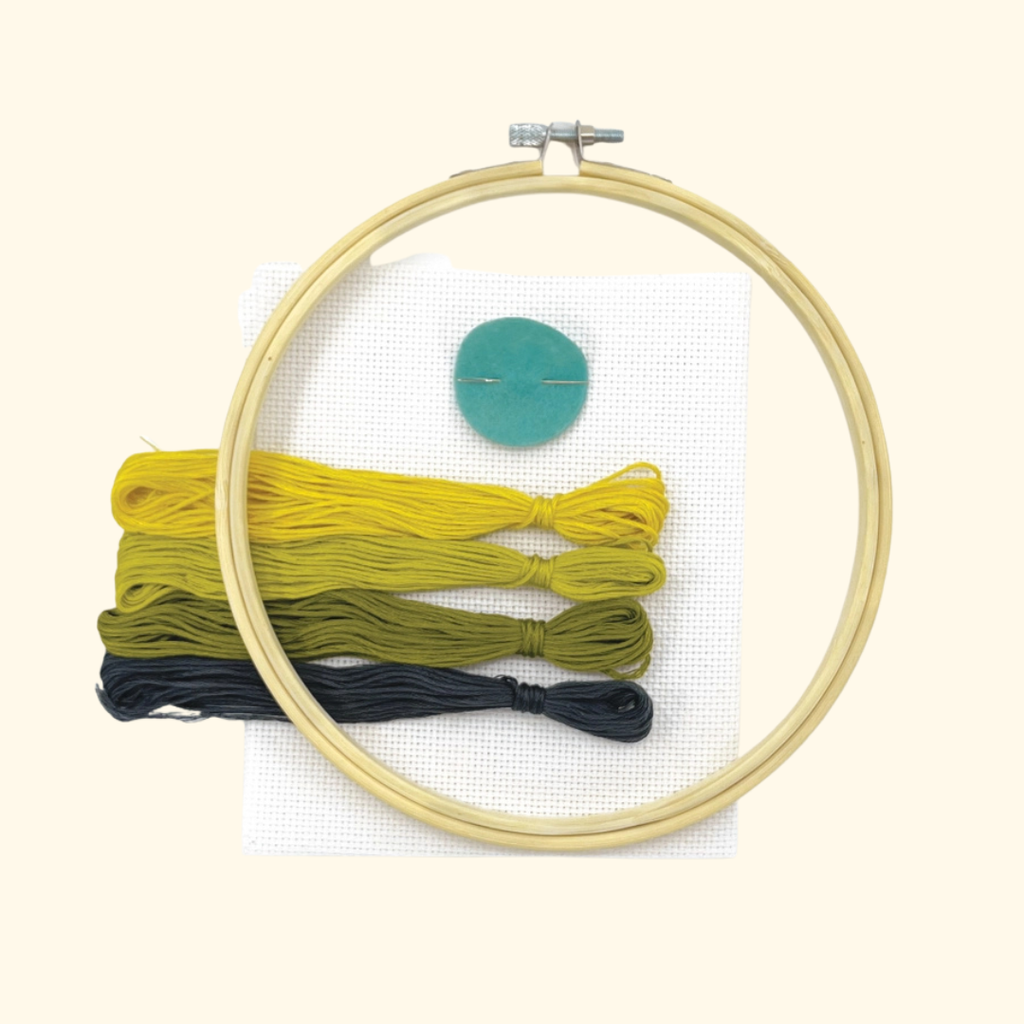 Embroidery kit with a wooden hoop, white canvas, three skeins of embroidery thread in yellow, olive green, and black, and a teal button on a light beige background.