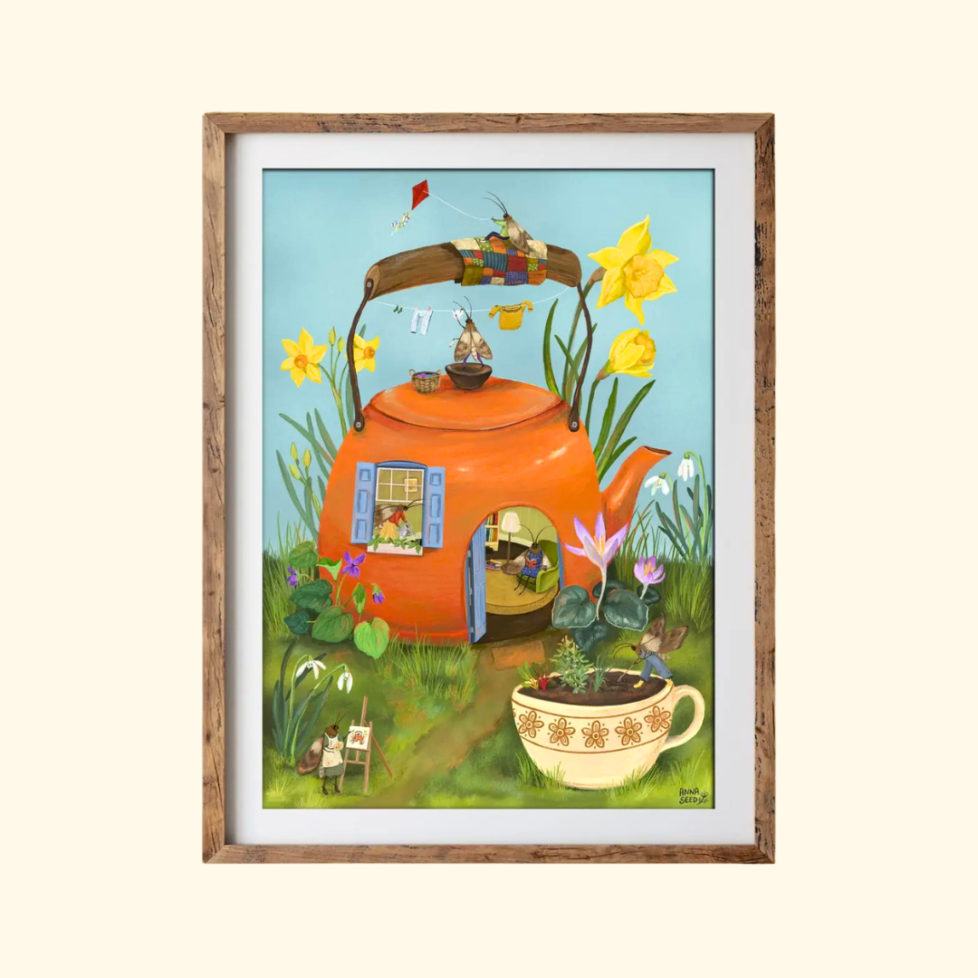 Teapot Cottage A4 art print by Anna Seed Art displayed in a timber frame, featuring a colourful teapot house in a whimsical garden setting.