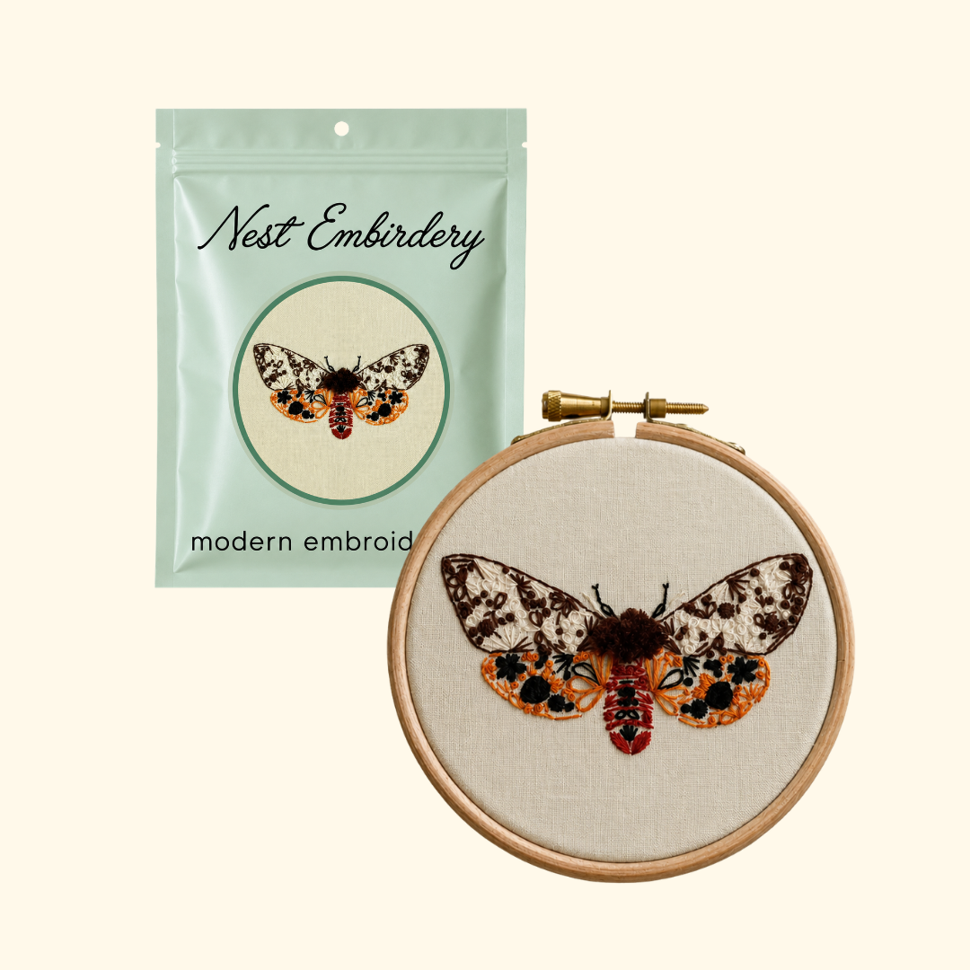 Nest Embirdery Tiger Moth Mini Embroidery Kit packaging displayed alongside the completed moth embroidery in its wooden hoop.
