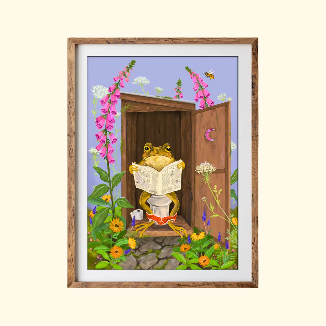 Toilet Toad A4 art print by Anna Seed Art displayed in a timber frame, featuring a toad reading on the toilet among colourful flowers and pink foxgloves.