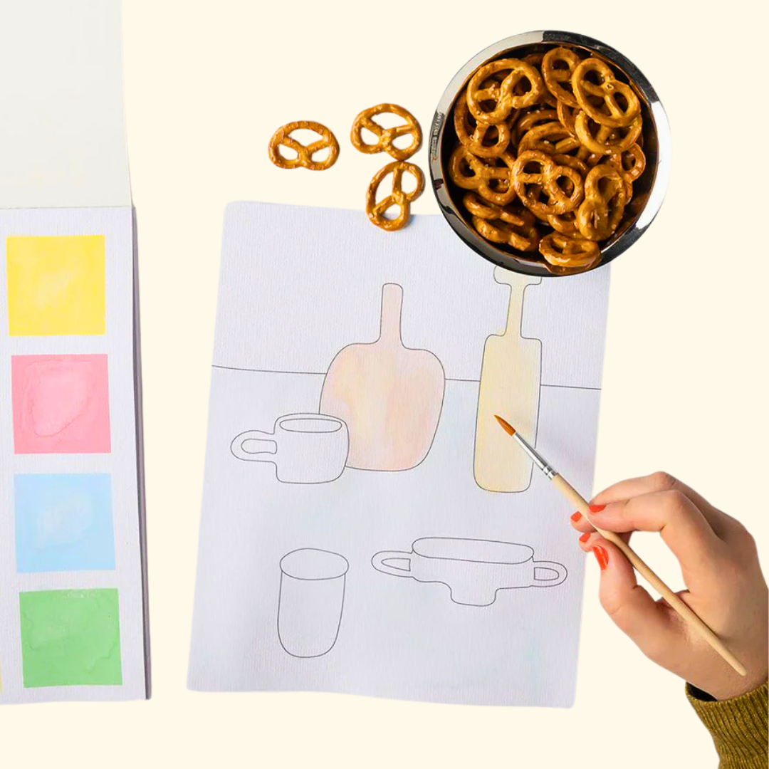 Painting a still life activity page from the Watercolour Painting Book beside a bowl of pretzels