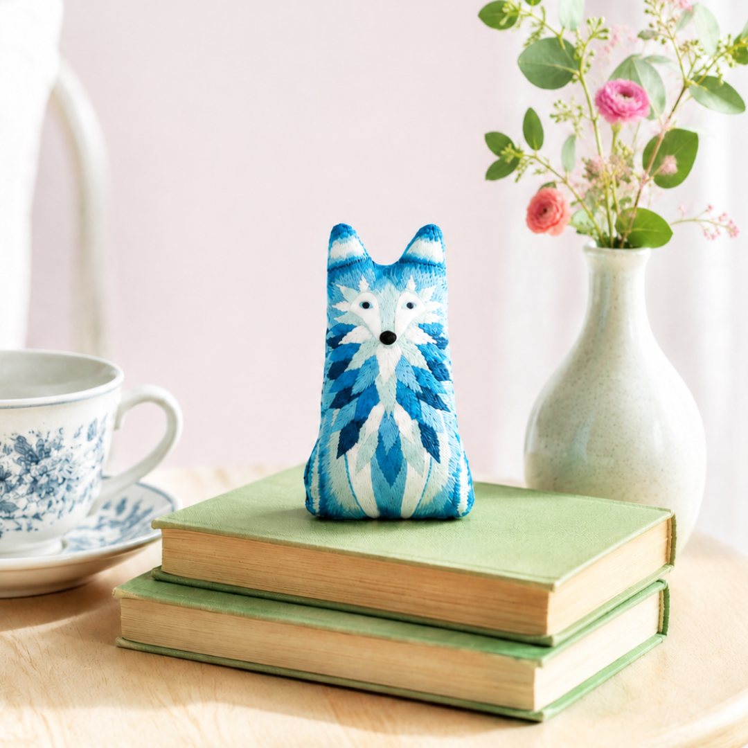 Finished Kiriki Press wolf embroidery doll displayed on stacked books beside flowers and a teacup.